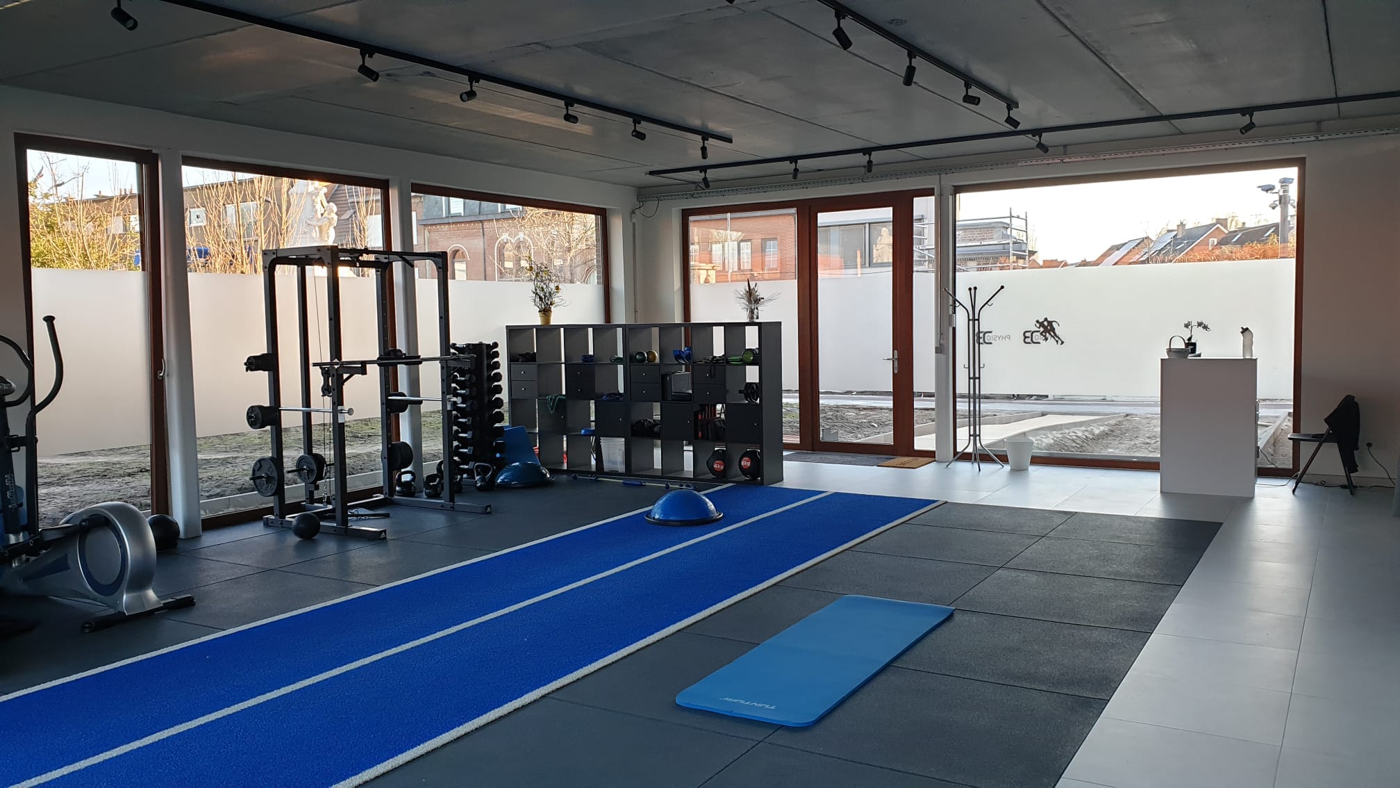 De praktijk van PhysioDB met een kast vol voorwerpen dat een kinesist gebruikt, een fitnes toestel en halters, en een indoor looptrack.
